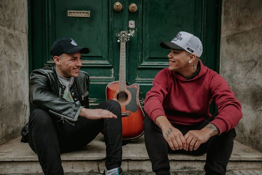 Zwei Musiker des Duos Chamo &amp; Dragger aus Argentinien und Uruguay, sitzend mit Gitarre in einer urbanen Szene – Symbol für die neue Generation lateinamerikanischer Musik, die traditionelle Klänge mit modernen Einflüssen verbindet.,
